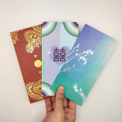 A hand holding three wedding hongbao (red envelopes) against a light background. The envelope on the left is red with gold dragon imagery, including stars and a moon. The middle envelope features a colorful design with a rainbow-like arch and a heart in the center, containing the double happiness symbol in purple. The right envelope has a gradient blue-green background with delicate white floral patterns. Each envelope showcases unique and intricate designs, reflecting traditional and modern aesthetics.