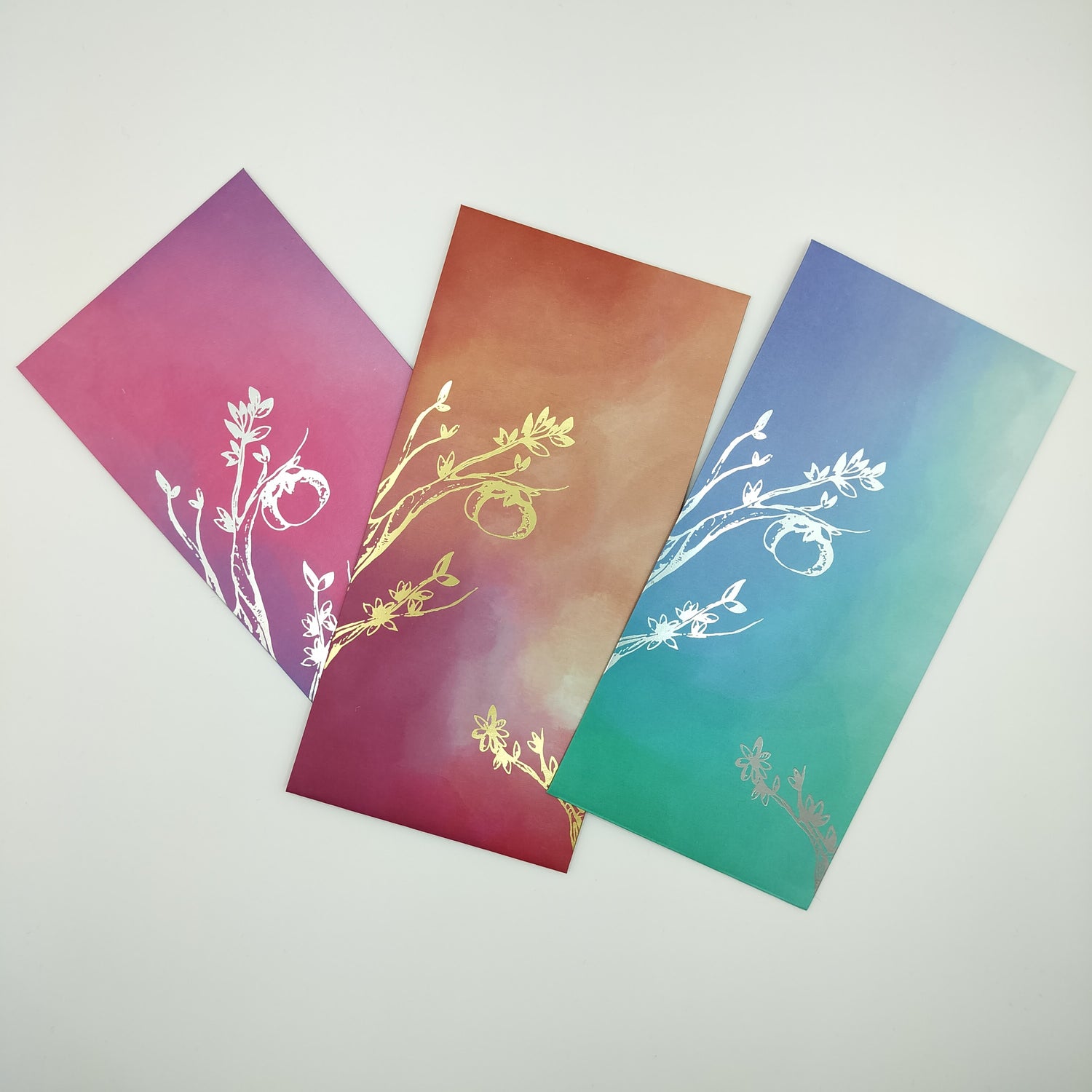 Three watercolour-style hong baos fanned out in an uneven horizontal line. The left is in the bi flag colours, the middle in the lesbian flag colours and the right in the gay flag colours. All have mandarin tree detailing in either gold or silver foil.